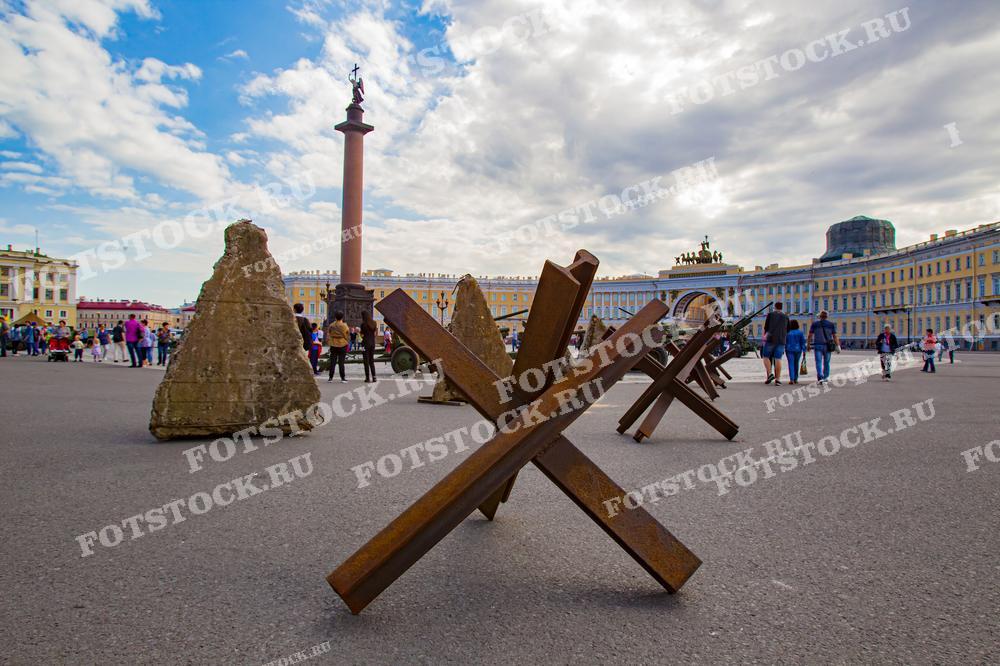 На фото запечатлена Дворцовая площадь в Санкт-Петербурге во время выставки вооружения времён Второй мировой войны. В центре кадра располагаются противотанковые ежи. Вдалеке видны Александровская колонна и Здание Главного штаба с его знаменитой триумфальной аркой. Это фото может быть использовано в учебных материалах по истории, в туристических брошюрах и руководствах по городу, а также в статьях о войне и блокаде.